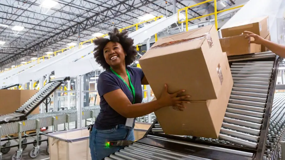 Una mujer sonriente recolectando cajas en la parte inferior de una manga de clasificación y empujándolas a lo largo de una cinta transportadora extensible hacia una estación de carga de camiones para su entrega