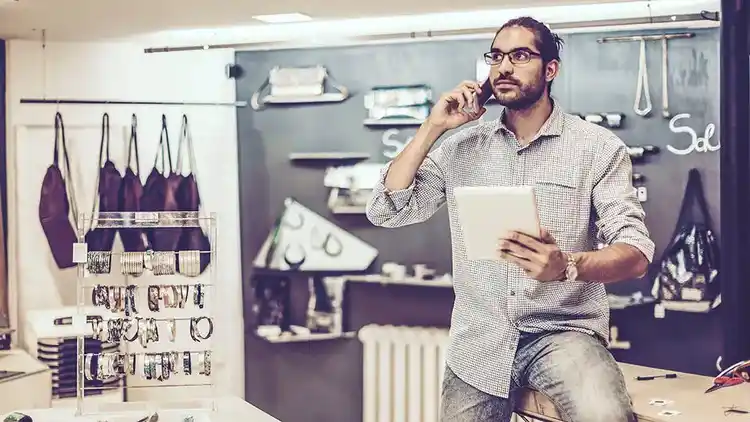 Man leans against desk while talking on a cell phone and holds a tablet