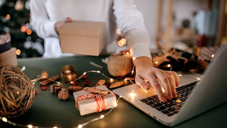 Close up of a desk with Christmas holiday lights and person’s hand on a laptop and holding a parcel.